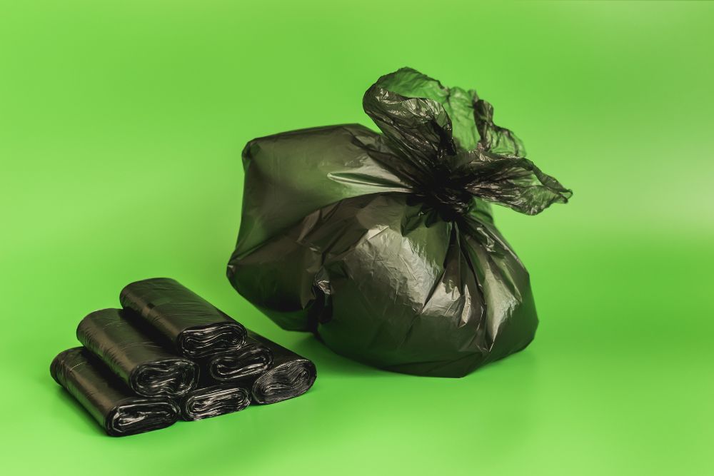 Bolsa de basura negra atada, rollos extra, fondo verde.